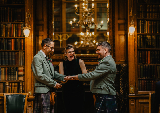 Photo of Linda, a middle-aged white woman, conducting a wedding ceremony for two men wearing kilts and tweed jackets. They are all laughing, and behind them is a glowing mantlepiece and library shelves